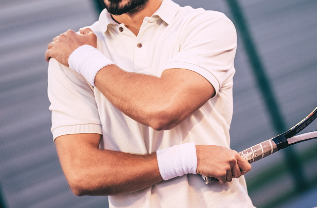 A man dressed in a collared shirt holds his shoulder in one hand and a tennis racket in the other.